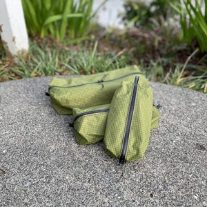 Peut inclure: Trois pochettes de rangement vert olive avec un motif &agrave; grille noire et des fermetures &eacute;clair noires. Les pochettes sont de forme rectangulaire et de tailles vari&eacute;es, fabriqu&eacute;es dans un tissu durable avec une texture visible et des tirettes.