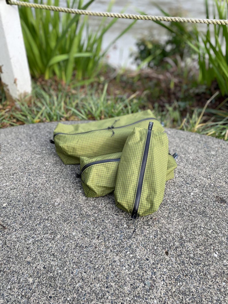 Peut inclure: Trois pochettes de rangement vert olive avec un motif &agrave; grille noire et des fermetures &eacute;clair noires. Les pochettes sont de forme rectangulaire et de tailles vari&eacute;es, fabriqu&eacute;es dans un tissu durable avec une texture visible et des tirettes.