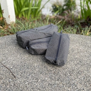 Peut inclure: Trois pochettes rectangulaires zipp&eacute;es de tailles diff&eacute;rentes, fabriqu&eacute;es dans un tissu noir avec un motif &agrave; carreaux blancs. Chaque pochette est dot&eacute;e d'une fermeture &eacute;clair noire sur le dessus pour ranger des petits objets en toute s&eacute;curit&eacute;.