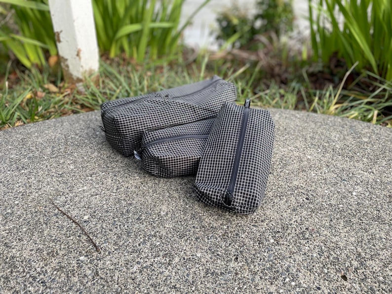 Peut inclure: Trois pochettes rectangulaires zipp&eacute;es de tailles diff&eacute;rentes, fabriqu&eacute;es dans un tissu noir avec un motif &agrave; carreaux blancs. Chaque pochette est dot&eacute;e d'une fermeture &eacute;clair noire sur le dessus pour ranger des petits objets en toute s&eacute;curit&eacute;.
