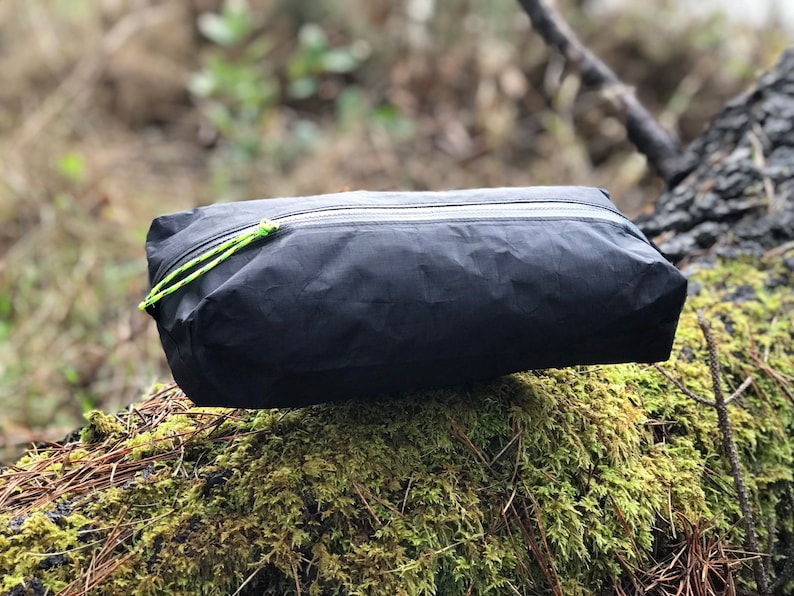 Peut inclure: Une pochette rectangulaire noire zipp&eacute;e fabriqu&eacute;e dans un mat&eacute;riau l&eacute;ger et froiss&eacute;. Le sac pr&eacute;sente une fermeture &eacute;clair horizontale sur le bord sup&eacute;rieur avec une tirette en cordon jaune n&eacute;on vif attach&eacute;e au curseur pour une manipulation ais&eacute;e.