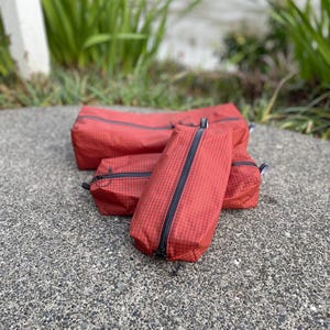 Peut inclure: Un ensemble de trois trousses rectangulaires de couleur rouge fonc&eacute; avec un motif &agrave; grille noire. Chaque trousse est dot&eacute;e d'une fermeture &eacute;clair noire le long du centre sup&eacute;rieur. Les trousses sont dispos&eacute;es en groupe, montrant des tailles vari&eacute;es.