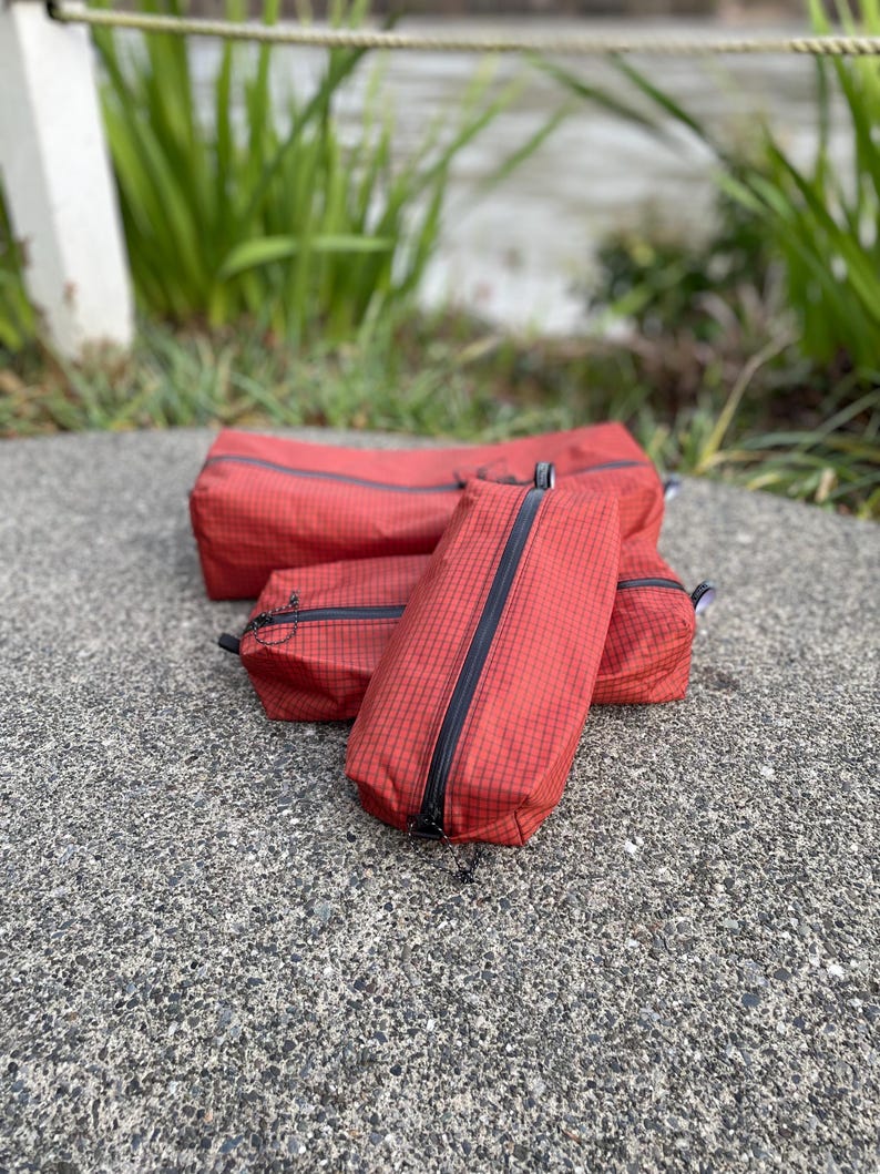Peut inclure: Un ensemble de trois trousses rectangulaires de couleur rouge fonc&eacute; avec un motif &agrave; grille noire. Chaque trousse est dot&eacute;e d'une fermeture &eacute;clair noire le long du centre sup&eacute;rieur. Les trousses sont dispos&eacute;es en groupe, montrant des tailles vari&eacute;es.