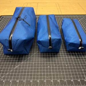 Peut inclure: Trois pochettes de rangement rectangulaires bleues de tailles diff&eacute;rentes, chacune dot&eacute;e d'une fermeture &eacute;clair noire r&eacute;sistante &agrave; l'eau au centre. Les pochettes sont fabriqu&eacute;es dans un mat&eacute;riau lisse et l&eacute;ger avec une forme structur&eacute;e.