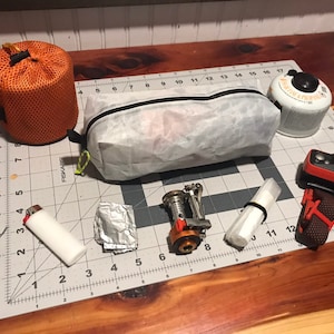 Peut inclure: Un ensemble d'&eacute;quipement de camping comprenant un sac en filet orange, une pochette zipp&eacute;e translucide blanche, une cartouche de gaz blanche, un briquet blanc, un morceau de papier aluminium, un r&eacute;chaud de camping en m&eacute;tal et une lampe frontale.
