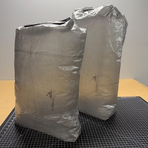 May include: Two silver-grey rectangular stuff sacks. The sacks are made of a lightweight, crinkled material. The text "ULTRALITESACKS" is printed on the front of each sack. The sacks are on a black cutting mat.