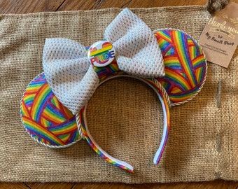 Op de afbeelding: Een Pride-hoofdband met regenboogkleurige oren en een grote, getextureerde witte strik. De oren hebben een kleurrijk regenboogpatroon en een knoop met het woord "PRIDE" is gecentreerd op de strik. De hoofdband is afgewerkt met gouden touw.