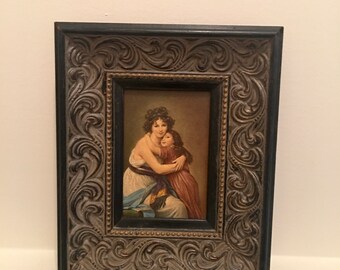 May include: A framed painting of a woman and a child embracing. The woman is wearing a white dress with a blue sash and the child is wearing a red dress. The painting is set in a dark wood frame with ornate carvings.