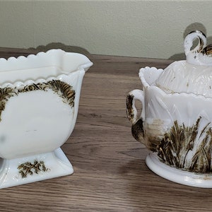 May include: A white milk glass creamer and sugar bowl set with brown and gold painted designs. The creamer has a handle and a scalloped rim. The sugar bowl has a lid with a swan finial.