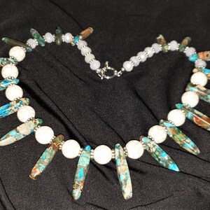 May include: A necklace with white and blue gemstone beads. The necklace features a long, pointed blue and brown gemstone pendant.