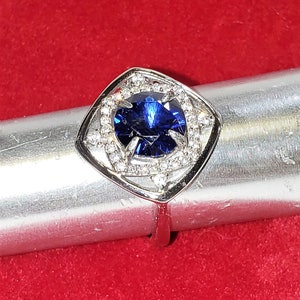 May include: A silver ring featuring a large, dark blue gemstone encircled by smaller, clear gemstones. The ring is set in a square-shaped design.