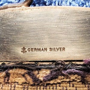 Peut inclure: Gros plan d'un objet argent&eacute; avec les mots "GERMAN SILVER" et un symbole d'ancre grav&eacute;s sur la surface.