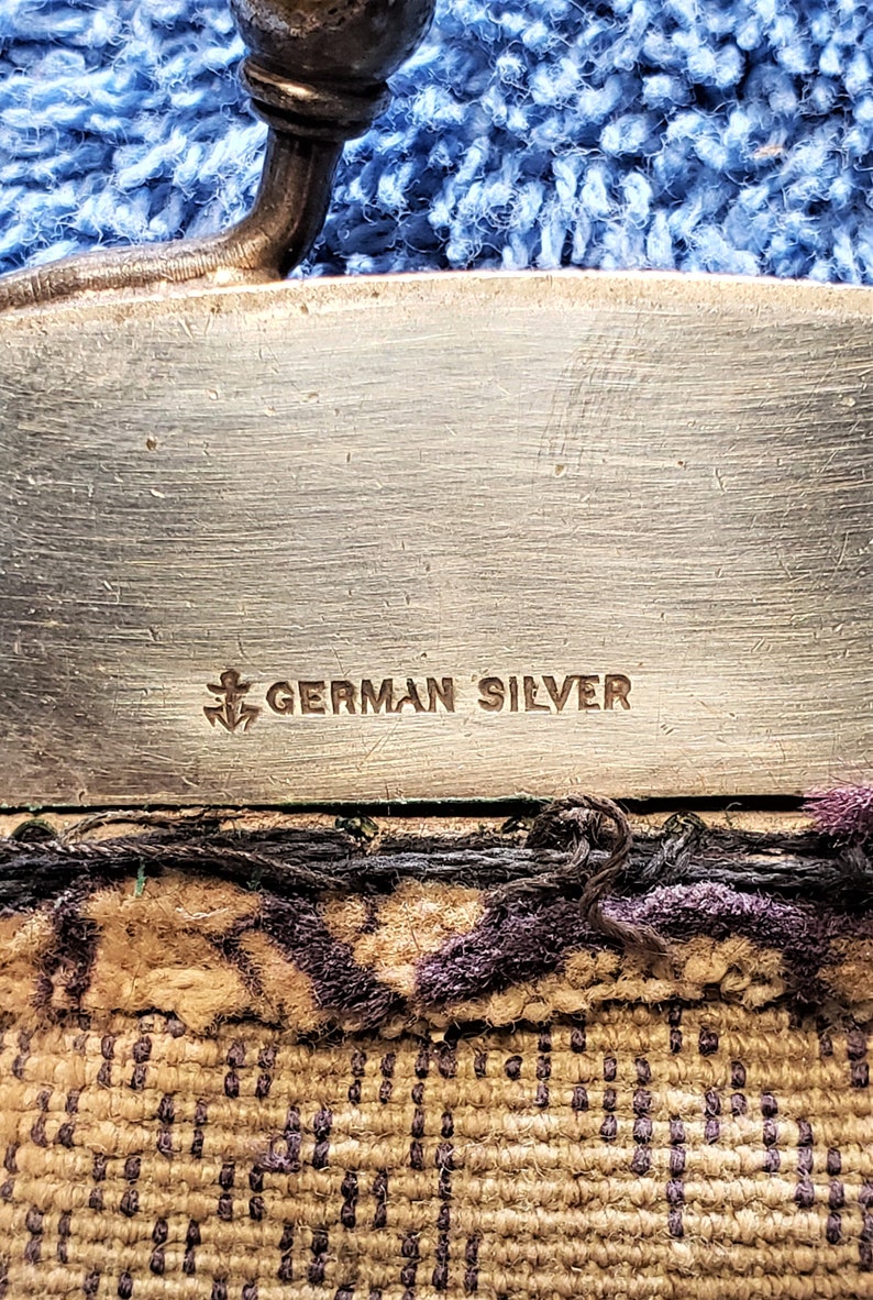 Peut inclure: Gros plan d'un objet argent&eacute; avec les mots "GERMAN SILVER" et un symbole d'ancre grav&eacute;s sur la surface.