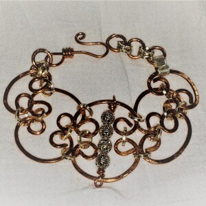 May include: A copper wire bracelet with a decorative design featuring multiple loops and circles. The bracelet has a clasp and a row of silver beads in the centre.