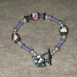 Könnte beinhalten: Ein lila-weißes Armband mit floralen Akzenten und einem silbernen Schiebeverschluss.