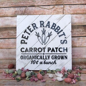 May include: White wooden sign with black lettering that reads "Peter Rabbit's Carrot Patch Organically Grown 10¢ a bunch". The sign has a black silhouette of three carrots in the center.