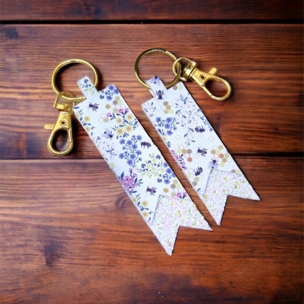 May include: Two white and gold keychains with a floral pattern and bee accents. The keychains are shaped like banners with a glittery finish.