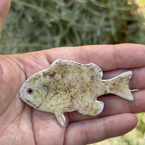 May include: A small, white ceramic fish figurine with brown and yellow paint details. The fish has a slightly curved body and a small, pointed tail.
