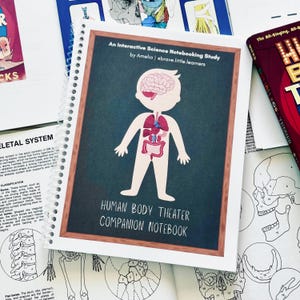 May include: A spiral-bound notebook titled Human Body Theater Companion Notebook by Amelia | ebrave.little.learners. The cover features a chalkboard-style frame with a central illustration of a human figure showing the brain and internal organs.
