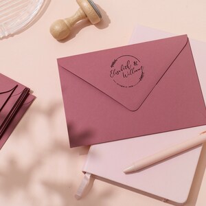 Op de afbeelding: Stofroze enveloppen met een cirkelvormige stempel met de namen "Elizabeth & William" en een datum. De enveloppen zijn gestapeld op een wit notitieboek met een pen. Een houten stempel en een glazen schaal zijn ook zichtbaar.