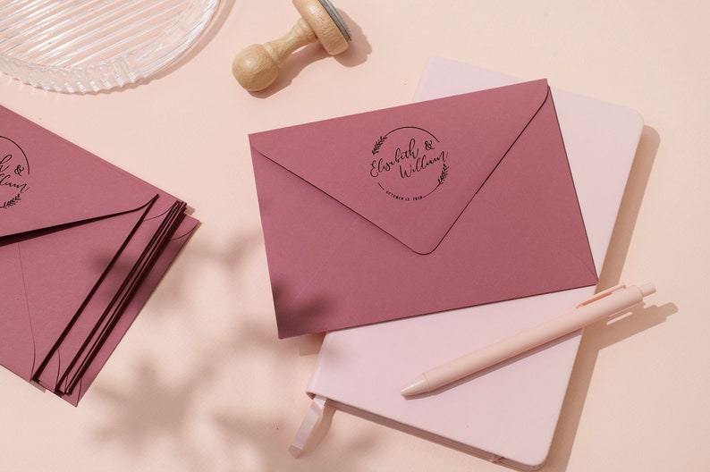 Op de afbeelding: Stofroze enveloppen met een cirkelvormige stempel met de namen "Elizabeth & William" en een datum. De enveloppen zijn gestapeld op een wit notitieboek met een pen. Een houten stempel en een glazen schaal zijn ook zichtbaar.