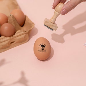 Puede incluir: Un huevo marr&oacute;n con un sello de goma negro que dice "Rosemary Farm, Free Range Eggs" sobre una superficie rosa. Una mano sostiene un sello de madera sobre el huevo.