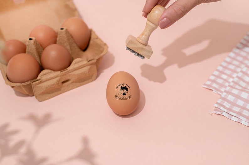 Puede incluir: Un huevo marr&oacute;n con un sello de goma negro que dice "Rosemary Farm, Free Range Eggs" sobre una superficie rosa. Una mano sostiene un sello de madera sobre el huevo.