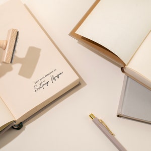 May include: An open journal with cream-colored pages, stamped with the text "THIS BOOK BELONGS TO Brittany Nguyen" in black ink. A wooden stamp is held above the page. A pen and other journals are also visible.