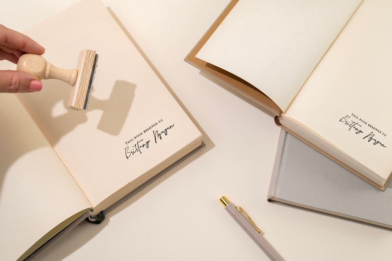 May include: An open journal with cream-colored pages, stamped with the text "THIS BOOK BELONGS TO Brittany Nguyen" in black ink. A wooden stamp is held above the page. A pen and other journals are also visible.