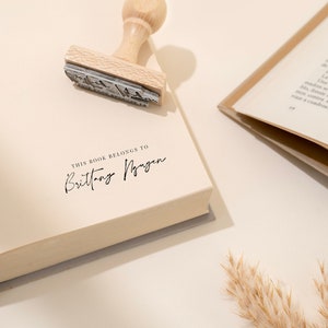 May include: A book with the text "THIS BOOK BELONGS TO Brittany Nguyen" stamped on the page. A wooden stamp is placed on the book. Another open book is in the background.