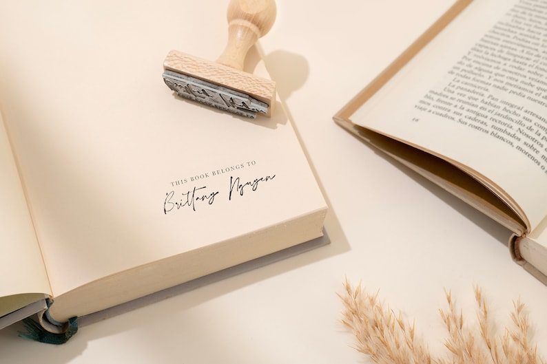 May include: A book with the text "THIS BOOK BELONGS TO Brittany Nguyen" stamped on the page. A wooden stamp is placed on the book. Another open book is in the background.