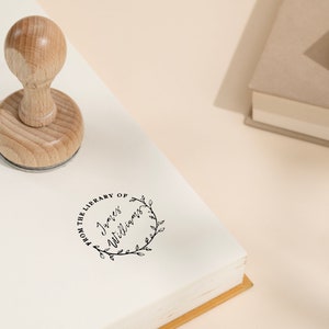 May include: A wooden library stamp with a round handle is pressed onto a book. The stamp imprint reads "FROM THE LIBRARY OF James Williams" within a circular floral wreath. The scene includes books and a pen on a light-colored surface.