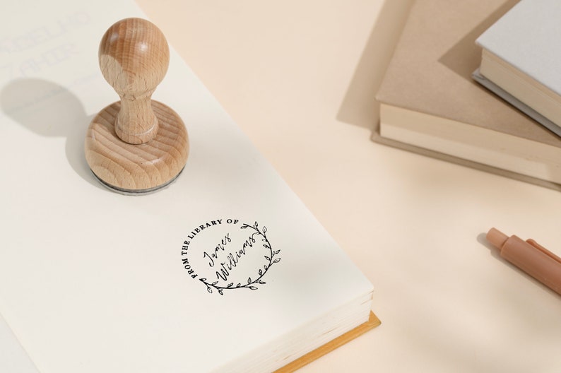 May include: A wooden library stamp with a round handle is pressed onto a book. The stamp imprint reads "FROM THE LIBRARY OF James Williams" within a circular floral wreath. The scene includes books and a pen on a light-colored surface.
