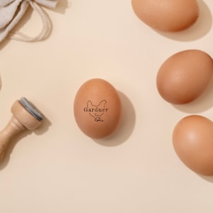 Puede incluir: Un huevo marr&oacute;n con un sello de goma que dice "Gardner Free Range" y una silueta de un pollo. El huevo est&aacute; sobre una superficie beige con otros huevos marrones.