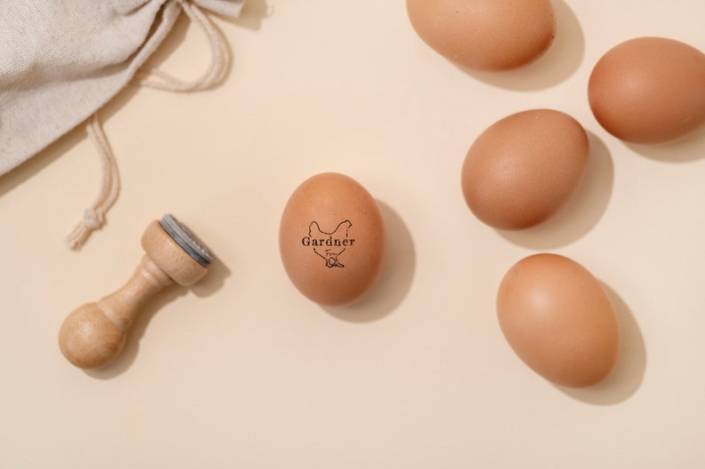 Puede incluir: Un huevo marr&oacute;n con un sello de goma que dice "Gardner Free Range" y una silueta de un pollo. El huevo est&aacute; sobre una superficie beige con otros huevos marrones.