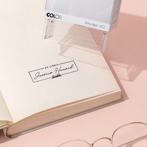 May include: A white and gray COLOP Printer 40 stamp is positioned over an open book. The stamp is creating a black ink imprint that reads "EX LIBRIS Jessica Howard" in a rectangular frame. The book's pages are cream-colored, and a pair of glasses rests below.