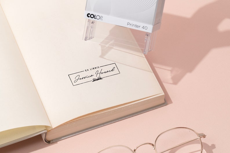 May include: A white and gray COLOP Printer 40 stamp is positioned over an open book. The stamp is creating a black ink imprint that reads "EX LIBRIS Jessica Howard" in a rectangular frame. The book's pages are cream-colored, and a pair of glasses rests below.