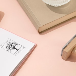 May include: An "Ex Libris" stamp with a tree design and the name "Patrick Lee" is next to a book. The stamp has a wooden handle and a rectangular stamping surface. Several books are visible, including a pink and a beige book, on a light pink surface.