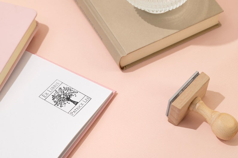 May include: An "Ex Libris" stamp with a tree design and the name "Patrick Lee" is next to a book. The stamp has a wooden handle and a rectangular stamping surface. Several books are visible, including a pink and a beige book, on a light pink surface.