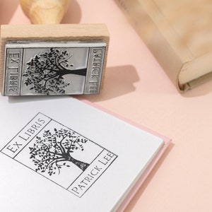 May include: A wooden stamp with a tree design and the text "EX LIBRIS PATRICK LEE" is pressed onto a white book. The stamp is next to a closed book with a beige cover. The background is a light pink.