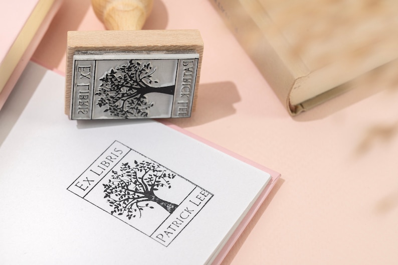 May include: A wooden stamp with a tree design and the text "EX LIBRIS PATRICK LEE" is pressed onto a white book. The stamp is next to a closed book with a beige cover. The background is a light pink.