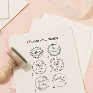 Op de afbeelding: Een houten stempel met een selectie van huwelijksontwerpen, waaronder bloemenkransen en namen. De tekst "Choose your design" staat bovenaan. Twee gouden ringen en briefpapier staan op de achtergrond.