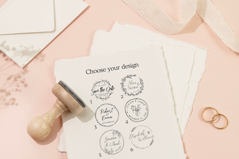 Op de afbeelding: Een houten stempel met een selectie van huwelijksontwerpen, waaronder bloemenkransen en namen. De tekst "Choose your design" staat bovenaan. Twee gouden ringen en briefpapier staan op de achtergrond.
