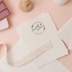 Op de afbeelding: Een verzameling trouwbriefpapier op een roze ondergrond. Inclusief cr&egrave;mekleurig papier met een gestempeld ontwerp, enveloppen en een decoratief lint. De stempel leest "Elisabeth & William" met een datum.