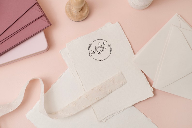 Op de afbeelding: Een verzameling trouwbriefpapier op een roze ondergrond. Inclusief cr&egrave;mekleurig papier met een gestempeld ontwerp, enveloppen en een decoratief lint. De stempel leest "Elisabeth & William" met een datum.