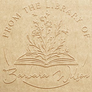 Puede incluir: Una superficie de papel color canela con un diseño circular en relieve. El texto dice FROM THE LIBRARY OF y Barbara Wilson. Una ilustración central muestra un libro abierto con flores silvestres y abejas.
