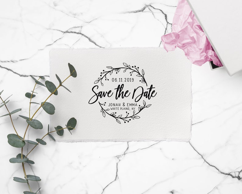 Op de afbeelding: Een witte kaart met de tekst "Save the Date" in sierlijk schrift, omringd door een bloemenkrans. De datum "06.11.2019" staat boven de tekst en de namen "Jonah & Emma, White Plains, NY" staan eronder. De kaart ligt op een marmeren oppervlak.