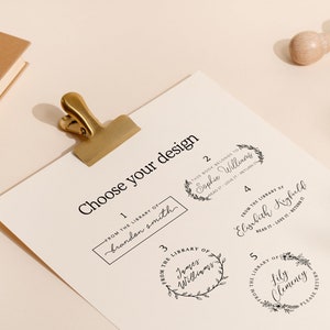 May include: A sheet of paper with the text "Choose your design" and five stamp design options. A gold clip holds the paper. A wooden stamp is at the top right. Books are in the top left.