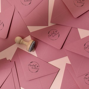 Op de afbeelding: Stofroze enveloppen met een ronde stempel met de tekst "Elisabeth & William" met een bloemenontwerp. Een houten stempel ligt naast de enveloppen. Voor huwelijksuitnodigingen of aankondigingen.