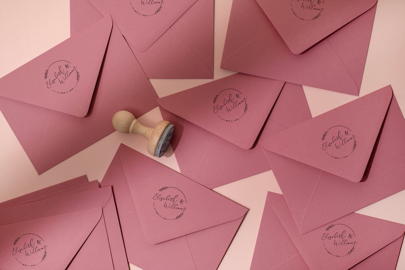 Op de afbeelding: Stofroze enveloppen met een ronde stempel met de tekst "Elisabeth & William" met een bloemenontwerp. Een houten stempel ligt naast de enveloppen. Voor huwelijksuitnodigingen of aankondigingen.